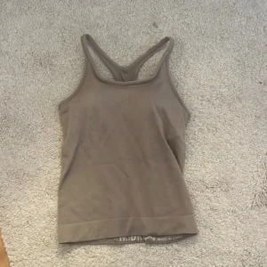 Beige ribbad linnetopp från TriDri - Säljer en beige ribbad tränings topp från TriDri. Toppen har en sportig design med brottarrygg och är tillverkad i ett stretchigt material som ger en bekväm passform. Perfekt för träning eller en avslappnad dag.