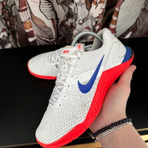 Nike träningsskor - Snygga träningsskor från Nike i vitt med en blå Swoosh och röd sula. Skorna har snörning och en modern design som passar perfekt för gymmet eller löprundan.