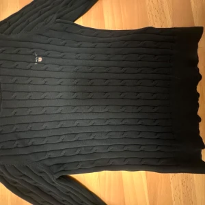 Svart stickad tröja från Gant - Säljer en stilren svart stickad tröja från Gant med kabelstickat mönster. Tröjan har långa ärmar och en rund halsringning. Perfekt för kyligare dagar och ger en klassisk look.
