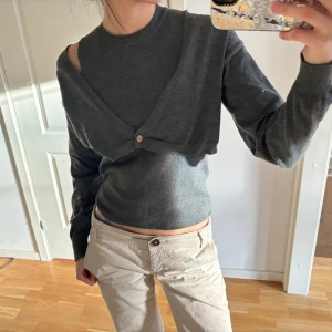 Grå stickad tröja med avtagbar bolerotopp - Säljer en stilren grå stickad tröja med en avtagbar bolerotopp. Tröjan är ärmlös och bolerotoppen har långa ärmar och knappar framtill. Perfekt för att variera din look!