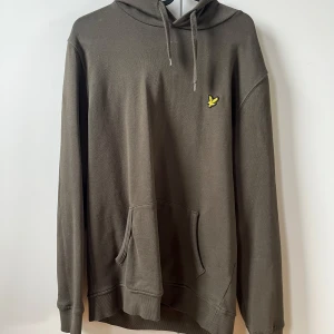 Grön hoodie från Lyle & Scott - Säljer en stilren grön hoodie från Lyle & Scott med en klassisk känguruficka och justerbar huva. Den har ett litet gult emblem på bröstet som ger en snygg detalj. Perfekt för en avslappnad look.