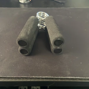 Svart handtränare med fjäder - Säljer en svart handtränare med mjuka skumhandtag och en robust metallfjäder. Perfekt för att stärka greppet och underarmarna. Lätt att använda hemma eller på kontoret.