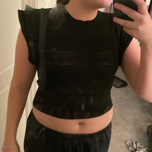 Svart topp från Zara - Säljer en svart topp från Zara med korta ärmar och volangdetaljer. Toppen är croppad och har ett diskret mönster i tygget.Toppen är även väldigt stretchig.