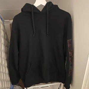 Svart hoodie med konstmotiv - Säljer en svart hoodie med en stor konstnärlig tryck på baksidan. Tröjan har en klassisk design med huva och dragsko samt en stor ficka framtill. Perfekt för dig som vill ha en unik stil med konstnärlig touch.