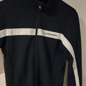 Svart träningströja från J.Lindeberg - Snygg mörkblå träningströja från J.Lindeberg med vita detaljer och dragkedja. Perfekt för sportiga aktiviteter. Jackan har en stilren design med långärmade ärmar och en hög krage.