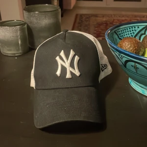 Svart och vit keps från New Era - Snygg svart och vit keps från New Era med ett broderat NY-logotyp framtill. Kepsen har en justerbar baksida i mesh för optimal passform och ventilation. Perfekt för en avslappnad stil.