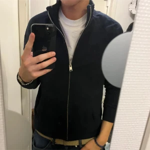 Svart tröja med dragkedja  - Säljer en stilren svart tröja från East West med dragkedja. Perfekt för en casual look. Tröjan har långa ärmar och en hög krage som kan bäras uppvikt eller nedvikt.