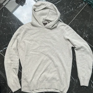 Beige hoodie från Oscar Jacobson - Säljer en stilren beige hoodie från Oscar Jacobson, tillverkad i Italien. Tröjan har en klassisk design med långa ärmar och en bekväm huva. Perfekt för en avslappnad look.