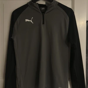 Grå och svart långärmad tröja från Puma Golf - Säljer en stilren grå och svart långärmad tröja från Puma med dragkedja vid halsen. Perfekt för träning eller vardagsbruk. Den har en sportig look med Pumas logga på bröstet. Tröjan har en liten fläck vid puma märket men inget som man tänker på 