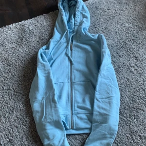 Ljusblå hoodie från H&M Divided - Säljer en ljusblå hoodie från H&M Divided. Den har en dragkedja framtill och en huva. Perfekt för en avslappnad stil. Hör av dig vid intresse! 