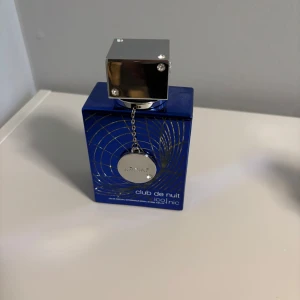 Armaf Club de Nuit blue Iconic Parfym - Säljer en elegant parfymflaska från Armaf, Club de Nuit Iconic. Flaskan är djupblå med ett stilrent silverlock och en dekorativ kedja. Perfekt för den som söker en sofistikerad doftupplevelse.