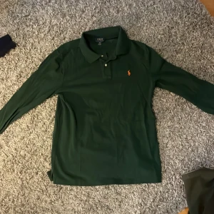 Grön pikétröja från Ralph Lauren - Säljer en snygg grön pikétröja från Ralph Lauren med klassisk krage och knappar. Tröjan har det ikoniska broderade logotypen i orange på bröstet. Perfekt för en stilren look! Nypris 2000