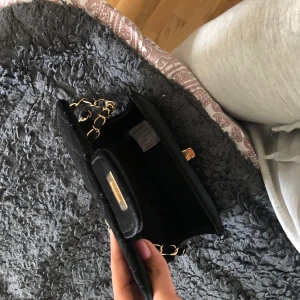 Svart axelväska med guldkedja - Säljer en svart axelväska med guldfärgad kedja och låsdetalj framtill. Perfekt storlek för att det viktigaste i väskan. Aldrig använt. Pris kan diskuteras