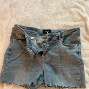 Blå jeansshorts med fransig kant från vero Moda  - Säljer ett par klassiska blå jeansshorts från vero moda med rå, fransig kant nertill. Shortsen har fem fickor, knappgylf och normal passform. Perfekta för varma dagar och enkel att matcha med allt.