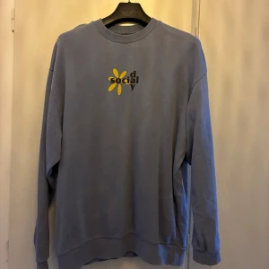 Ljusblå sweatshirt med tryck  - Ljusblå sweatshirt med stort svart och gult tryck. Tröjan har rund hals och långa ärmar. Perfekt för en avslappnad stil. Strl S (oversized) 