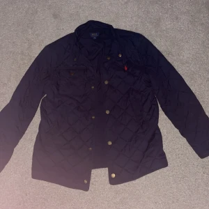 Mörkblå quiltad jacka från Polo Ralph Lauren - Säljer en mörkblå quiltad jacka från Polo Ralph Lauren med guldiga knappar och klassisk röd logga på bröstet. Jackan har lång ärm, två bröstfickor och en stilren look som passar året runt