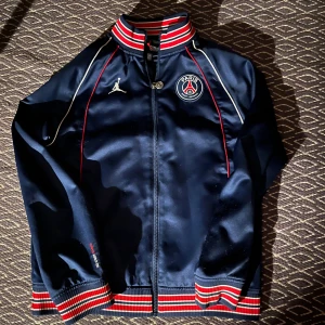 Mörkblå PSG x Jordan track jacket - Säljer en mörkblå track jacket från Jordan och PSG med broderad logga på bröstet och stor 'PARIS' text på ryggen. Jackan har dragkedja, randiga muddar i rött, vitt och blått samt tunna röda detaljer längs sömmarna. Den är S men funkar som M.