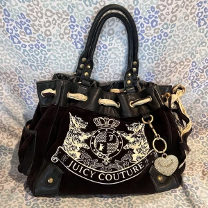 Svart axelväska från Juicy Couture med emblem - Juicy Couture axelväska i svart sammet med stora handtag och dekorativt vitt emblem framtill. Väskan har snörning upptill, detaljer i skinn. Perfekt för dig som gillar statement-accessoarer. Fast pris (2300)