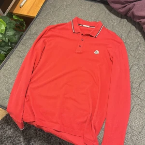 Röd långärmad pikétröja från Moncler - Säljer en röd långärmad pikétröja från Moncler med klassisk krage och knappar framtill. Tröjan har Moncler-logga på bröstet och vit-svart rand på kragen. Skön inför sommaren. Taggen i loggan på bröstet fungerar som den ska. 
