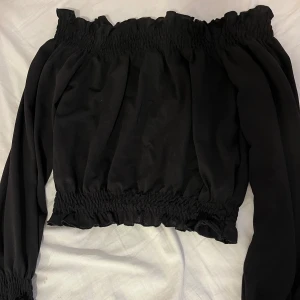 Svart offshoulder topp med volang - Trendig svart offshoulder topp med volangdetaljer vid halsringning, midja och ärmar. Resår upptill och nedtill för en bekväm passform.