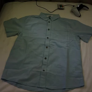 Ljusblå kortärmad skjorta från H&M - Säljer en ljusblå skjorta från H&M med korta ärmar och klassisk krage. Skjortan har knappar framtill och en bröstficka på vänster sida. Perfekt för varma dagar och enkel att matcha med olika outfits.