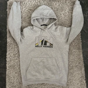 Grå hoodie från Helly Hansen - Säljer en grå hoodie från Helly Hansen med tryckt logga på bröstet i svart, vitt och gult. Tröjan har huva med dragsko och en stor magficka framtill. Perfekt för en avslappnad stil.