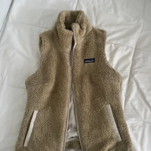 Beige fleeceväst från Patagonia - Mysig beige fleeceväst från Patagonia med hög krage och dragkedja framtill. Västen har två fickor med vita detaljer och är perfekt för lager-på-lager-stil. Klassisk logga på bröstet.