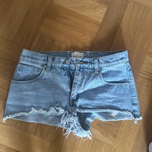 Ljusblå jeansshorts med fransar - Säljer ett par ljusblå jeansshorts från Armedangels med råa franskantade benslut och klassisk femficksmodell. Shortsen har normal passform och låg midja, perfekta för sommardagar.