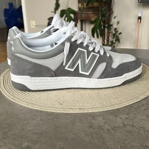 Grå New Balance sneakers - Snygga grå och vita New Balance sneakers med klassisk design och stora N-loggor på sidorna. Skorna har ovandel i mocka och syntet, vita snören och platt sula. Perfekta för en stilren och sportig look.