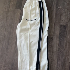 Vita byxor - Säljer ett par vita byxor med svarta ränder längs sidorna och logga på låret. Byxorna har elastisk midja och en avslappnad passform, perfekta för en sportig look.