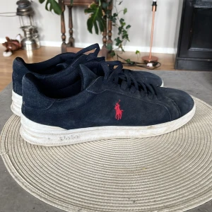 Svarta sneakers från Polo Ralph Lauren - Svarta sneakers i mocka från Polo Ralph Lauren med vit sula och röd broderad logga på sidan. Klassisk rund tå och snörning framtill. Perfekta för en stilren och avslappnad look.