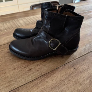 Svarta boots från Fiorentini + Baker - Säljer ett par svarta boots från Fiorentini + Baker i skinn med rund tå och dekorativt spänne i metall på sidan. Klassisk modell med markerade sömmar och robust sula. Perfekta för dig som gillar tidlös stil och kvalitet.