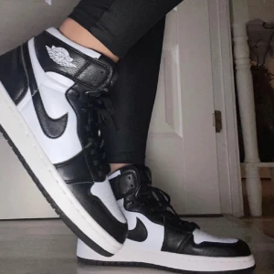 Nike Air Jordan 1 svartvita sneakers - Säljer ett par klassiska Nike Air Jordan 1 sneakers i svart och vitt. Skorna har högt skaft, svart snörning och den ikoniska swoosh-loggan på sidan. Perfekta för dig som gillar streetstyle och vill ha en tidlös look.