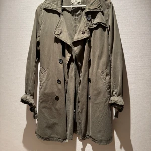 Olivgrön trenchcoat från Maison Scotch - Snygg olivgrön trenchcoat från Maison Scotch med klassisk krage och knappar framtill. Jackan har långa ärmar, fickor på sidorna och ett avslappnat snitt. Perfekt för lager-på-lager under vår och höst.