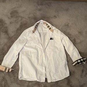 Vit skjorta med klassiskt rutigt mönster från Burberry - Säljer en vit skjorta från Burberry med deras ikoniska beige, svarta och röda rutiga detaljer på insidan av kragen och ärmsluten. Skjortan har lång ärm, knappar framtill och en broderad logga på bröstet.