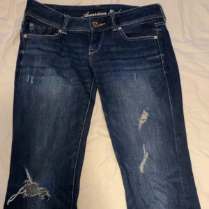 Blå slitna jeans från American Eagle - Mörkblå jeans från American Eagle med slitningar och hål på benen. Klassisk femficksmodell med vita kontrastsömmar och knappgylf. Jeansen har en boot passform och en låg midja. Inga sakador utan alla slitningar är till för att vara där! Mått: midja rakt över - 38cm innerbenslängd - 80cm.