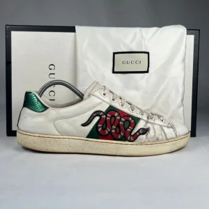 Gucci Ace sneakers med orm brodyr - Gucci Ace sneakers i vitt skinn med ikonisk röd och grön orm brodyr på sidan. Skorna har gröna och röda detaljer på hälen, snörning framtill och platt sula. Klassisk rund tå och diskret Gucci-logga på baksidan.