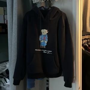 Mörkblå hoodie med björntryck från Polo Ralph Lauren - Svart hoodie från Polo Ralph Lauren med en söt björn i jeansjacka och röd tröja på bröstet. Klassisk huva och känguruficka framtill. Perfekt för en avslappnad och cool stil. Den e ganska liten för att vara M.