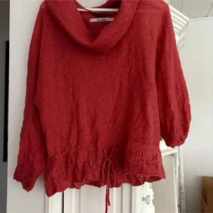 Röd stickad tröja med volang  - Mysig röd stickad tröja från RoseBud med bred krage och fina volangdetaljer samt knytning nertill. Perfekt för kyligare dagar och enkel att styla till olika outfits.