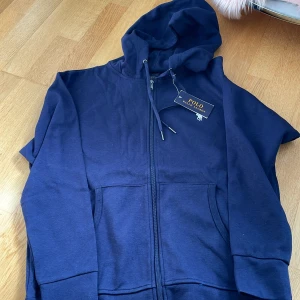 Mörkblå hoodie från Polo Ralph Lauren - Säljer en mörkblå hoodie från Polo Ralph Lauren med dragkedja och huva. Tröjan har fickor framtill och den klassiska broderade loggan i vitt på bröstet. Perfekt för en avslappnad stil.
