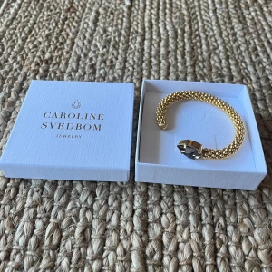 Guldfärgat armband med stor kristall från Caroline Svedbom - Helt oanvänd! Säljer ett exklusivt guldfärgat armband från Caroline Svedbom med en stor, droppformad kristall i färgen greige. Armbandet har en elegant och lyxig design som verkligen sticker ut. Nypris 995kr. 