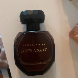 Rebecca Stella Date Night Eau de Parfum - En elegant parfymflaska i mörkbrunt glas med svart, multifasetterat lock. 'Date Night' från Rebecca Stella. Texten är i guld på framsidan. Nypris var 700. 50 ml med mer än 2/3 kvar. 