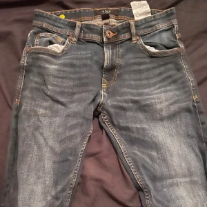 Blå jeansbyxor med slitningar - Säljer ett par klassiska blå jeansbyxor med snygga slitningar och kontrastsömmar. Byxorna har fem fickor, knappgylf och raka ben. Perfekta för en avslappnad stil. Märke Smog