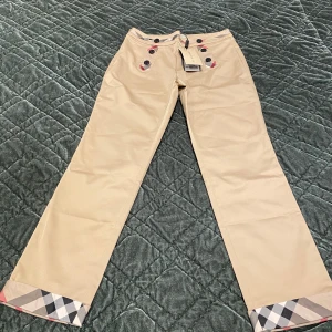 Beige byxor från Burberry med rutiga detaljer - Snygga beige byxor från Burberry med klassiska rutiga detaljer vid benslut och knappar framtill. Byxorna har normal passform och är perfekta för dig som gillar stilrena plagg med lyxig känsla. Ikoniskt Burberry-mönster på insidan av linningen och vid knappar.