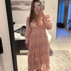 Långklänning i blommigt mönster med volang - Säljer en lång balklänning i ljusa rosa och orange toner med blommigt mönster. Klänningen har långa ärmar med knappar vid ärmslut, v-ringning och volangdetaljer nertill. Perfekt för sommarens tillfällen!