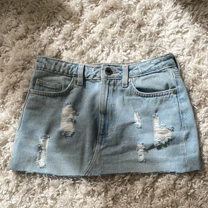 Ljusblå jeanskjol med slitningar - Säljer en kort lågmidjadljusblå jeanskjol med flera slitna detaljer framtill. Kjolen har klassisk femficksmodell och knappstängning i midjan. Perfekt för dig som gillar en avslappnad och trendig stil. Midjemåttet är 35 cm🫶💕