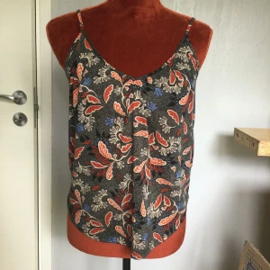 Blommig linnetopp från H&M Conscious - Säljer en tunn och luftig linnetopp från H&M Conscious i storlek XS. Toppen har smala axelband och ett färgglatt blommigt mönster i rött, blått och beige mot svart bakgrund. Perfekt för varma dagar! Mönstrat Linne i polyester  Längd: 55 cm Armhåla till armhåla: 47cm Katter finns i hemmet!