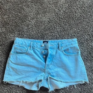 Ljusblå jeansshorts från 157 - Ljusblå jeansshorts från 157 med råa fransar nertill och klassisk femficksdesign. Knappar framtill och bälteshällor. Perfekta för varma dagar och enkel att matcha med olika toppar.