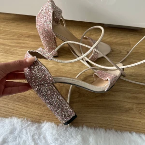 Glittriga rosa klackskor med rosett - Säljer ett par rosa glittriga klackskor med bred klack och öppen tå. Skorna har en söt rosett bak och tunna remmar som knyts runt vristen. Perfekta för att sticka ut på festen! Oanvända storlek 36