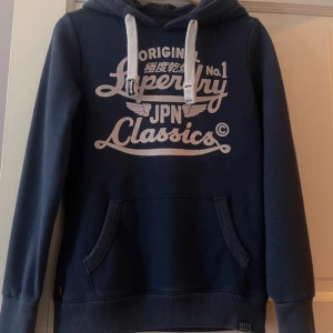 Superdry hoodie - Mörkblå hoodie från Superdry med vit tryckt logga och text på bröstet. Tröjan har huva med vita snören och en stor magficka framtill. Perfekt för en avslappnad stil och enkel att matcha med jeans eller joggers.